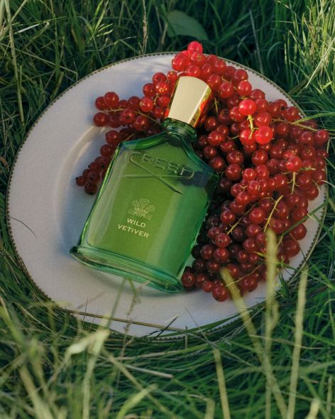 CREED Wild Vetiver 岩蘭野境香水瓶置於白色瓷盤與紅色果實間的草地場景